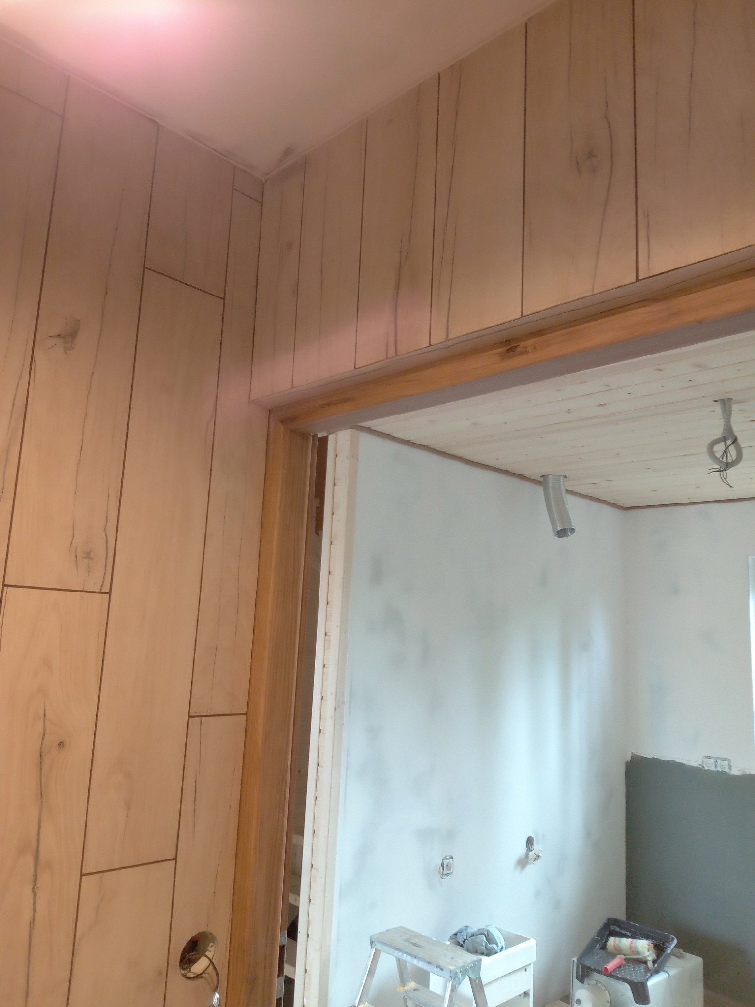 Ściana wykończona panelami imitującymi drewno, sąsiadująca z pomalowaną na biało ścianą w trakcie prac przygotowawczych w Narolu. Widoczna drabina i narzędzia malarskie.