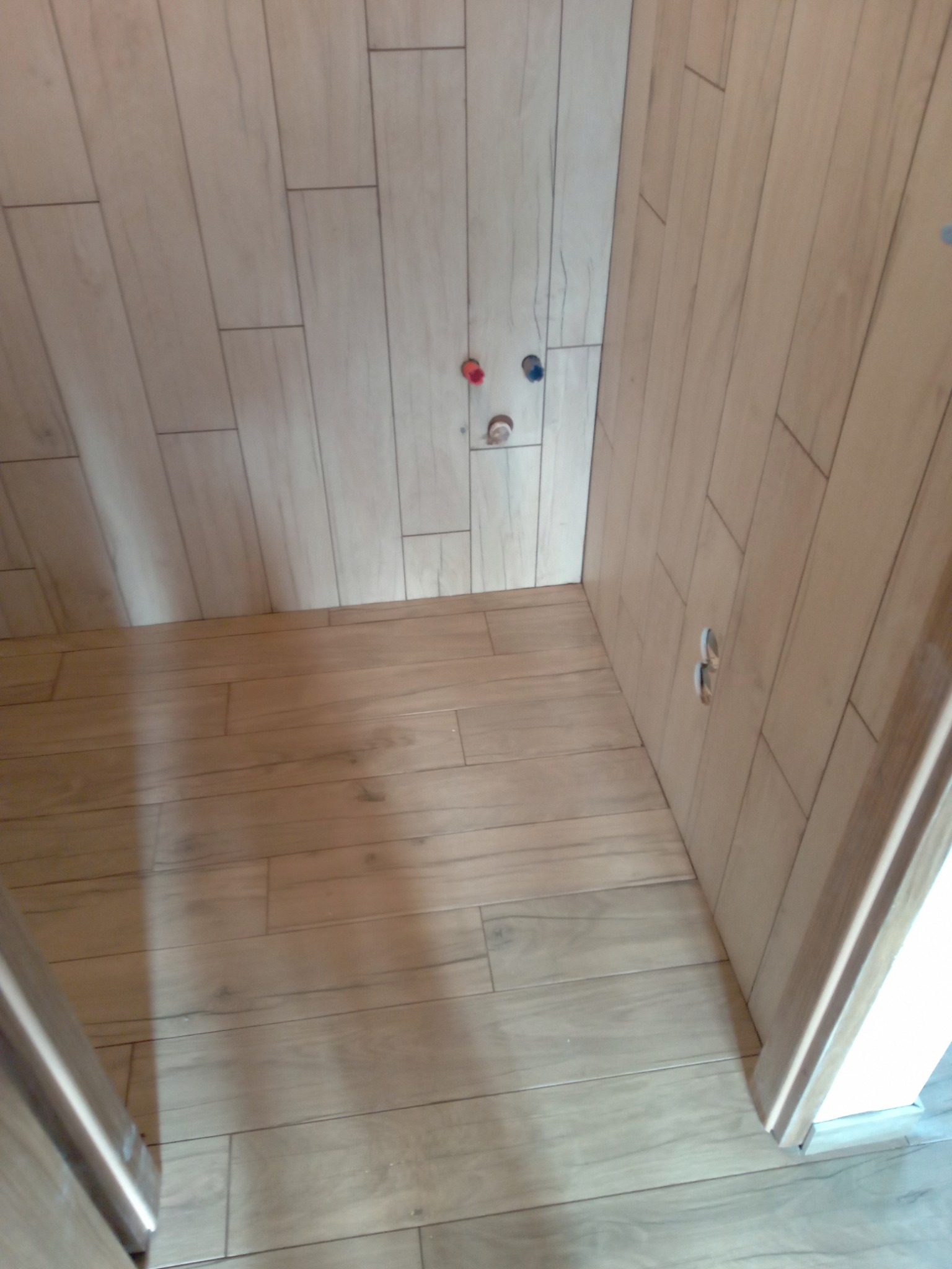 Ściany i podłoga wyłożone płytkami imitującymi drewno w trakcie prac wykończeniowych; widoczne wystające rury instalacji wodnej i otwór na gniazdko elektryczne.
