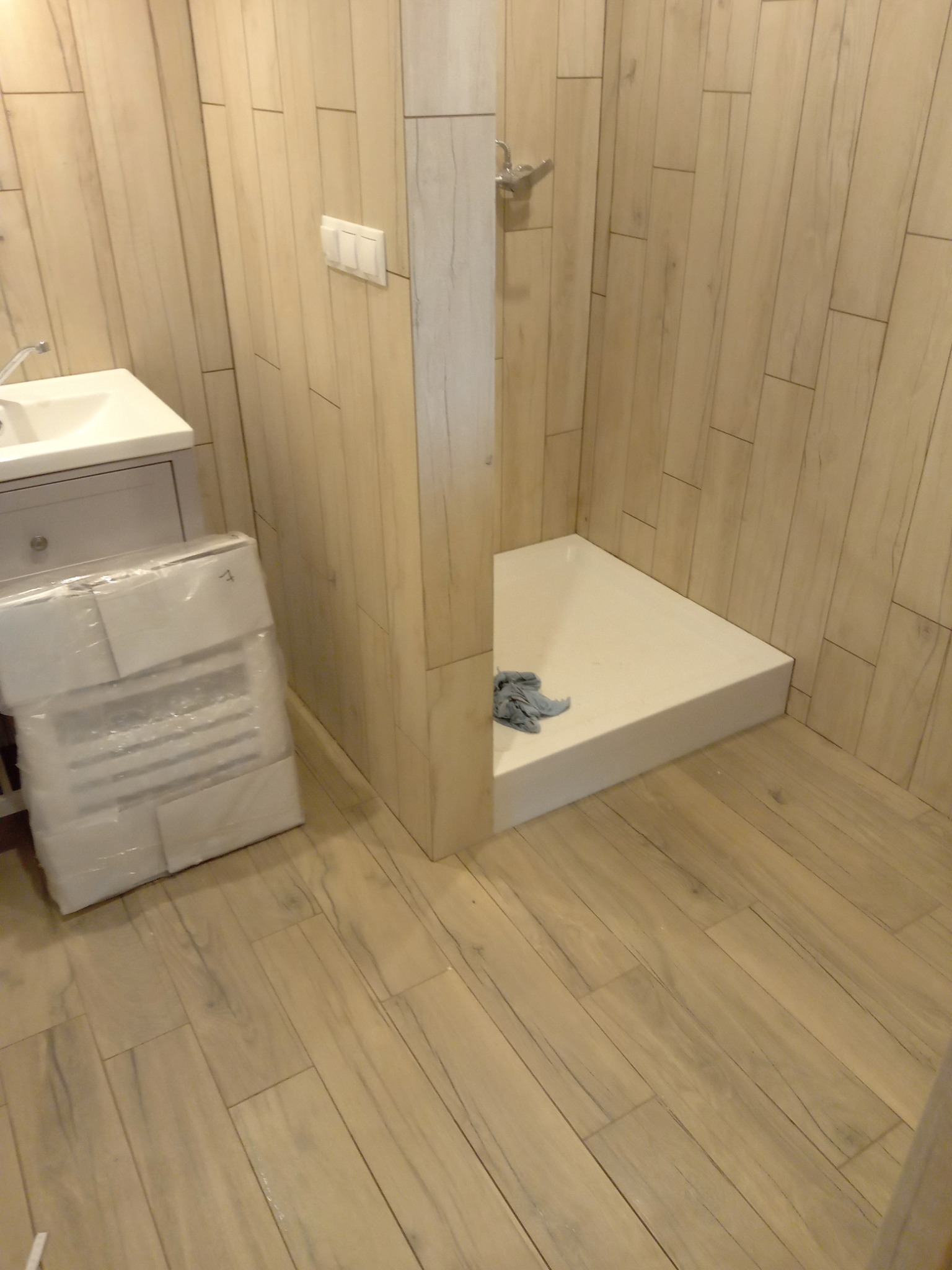 Narożna kabina prysznicowa z niskim brodzikiem, obok umywalka z szafką i zapakowany element wyposażenia, wykończona płytkami imitującymi drewno, widoczny fragment szmatki w brodziku.