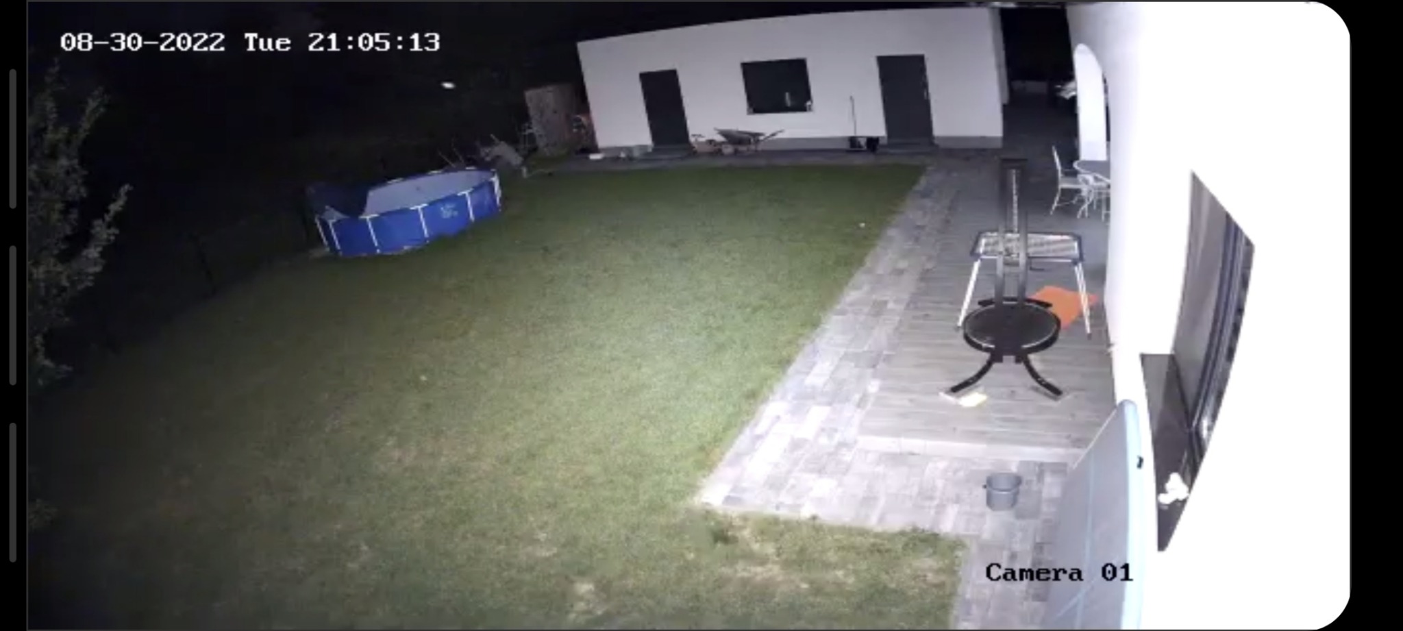 Obraz z kamery monitoringu nocą, widok na trawnik, basen, budynek gospodarczy z trzema wejściami oraz elementy wyposażenia ogrodu.
