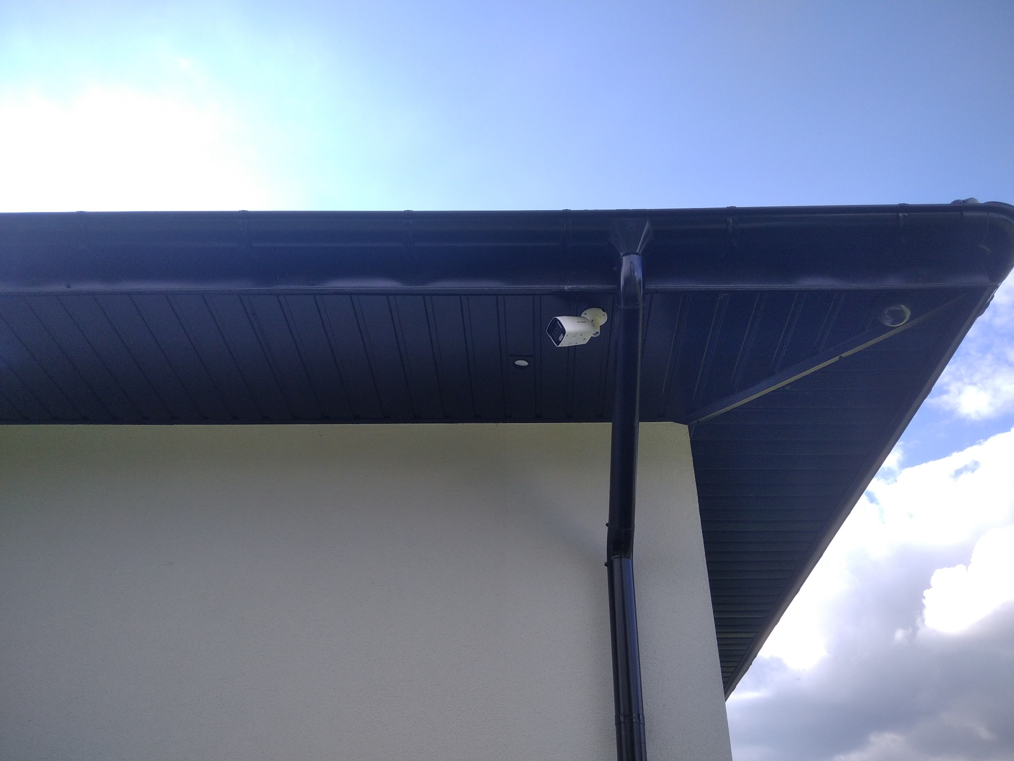 Kamera monitoringu zamontowana pod okapem budynku z ciemną blachodachówką i rynnami, widok z dołu na tle jasnego nieba.
