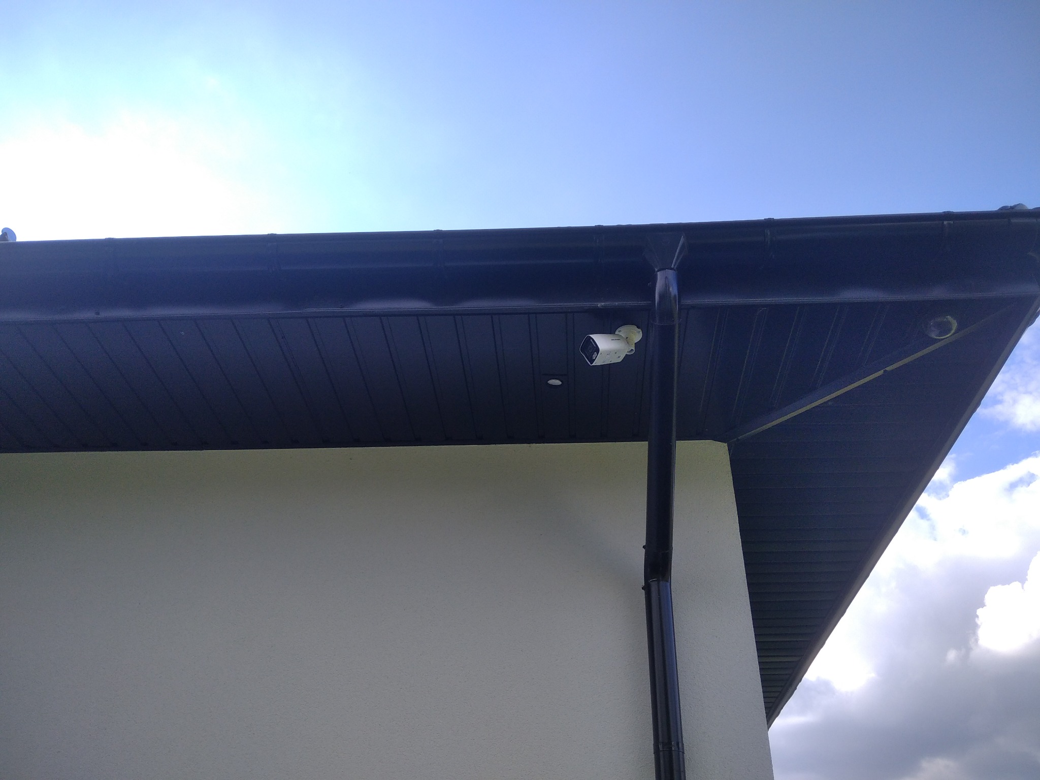 Biała kamera monitoringu zamontowana pod ciemnym okapem domu, widok z dołu na tle błękitnego nieba.