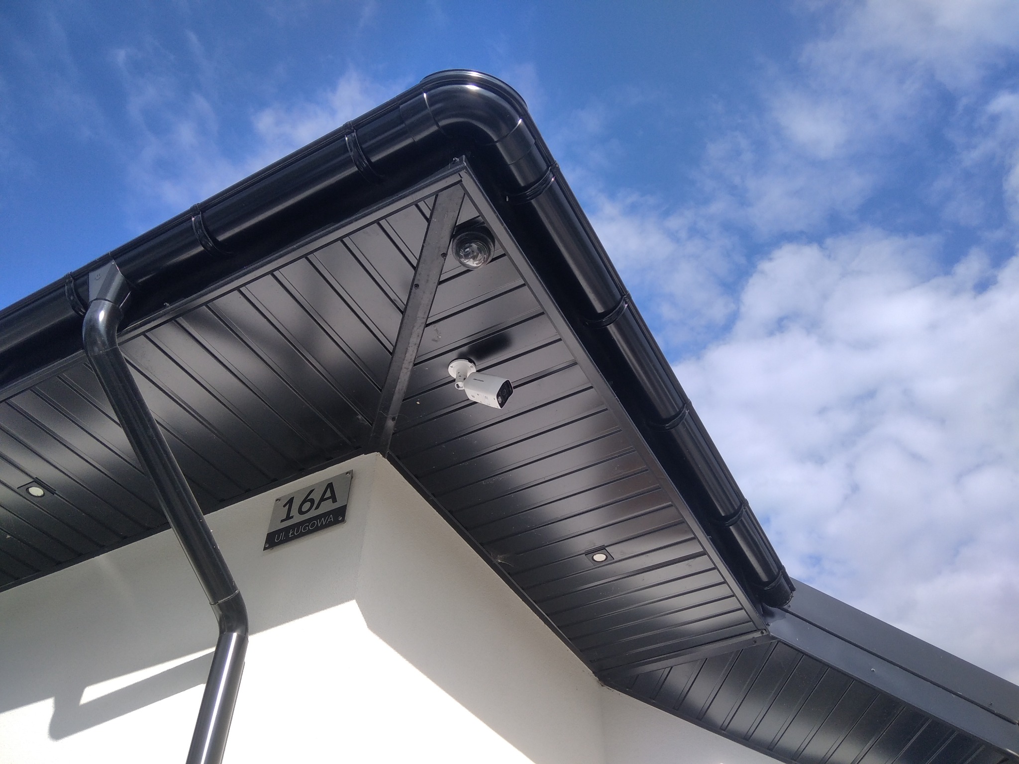 Dwie kamery monitoringu zamontowane pod czarnym, metalowym zadaszeniem budynku z adresem 16A na tle błękitnego nieba z chmurami.