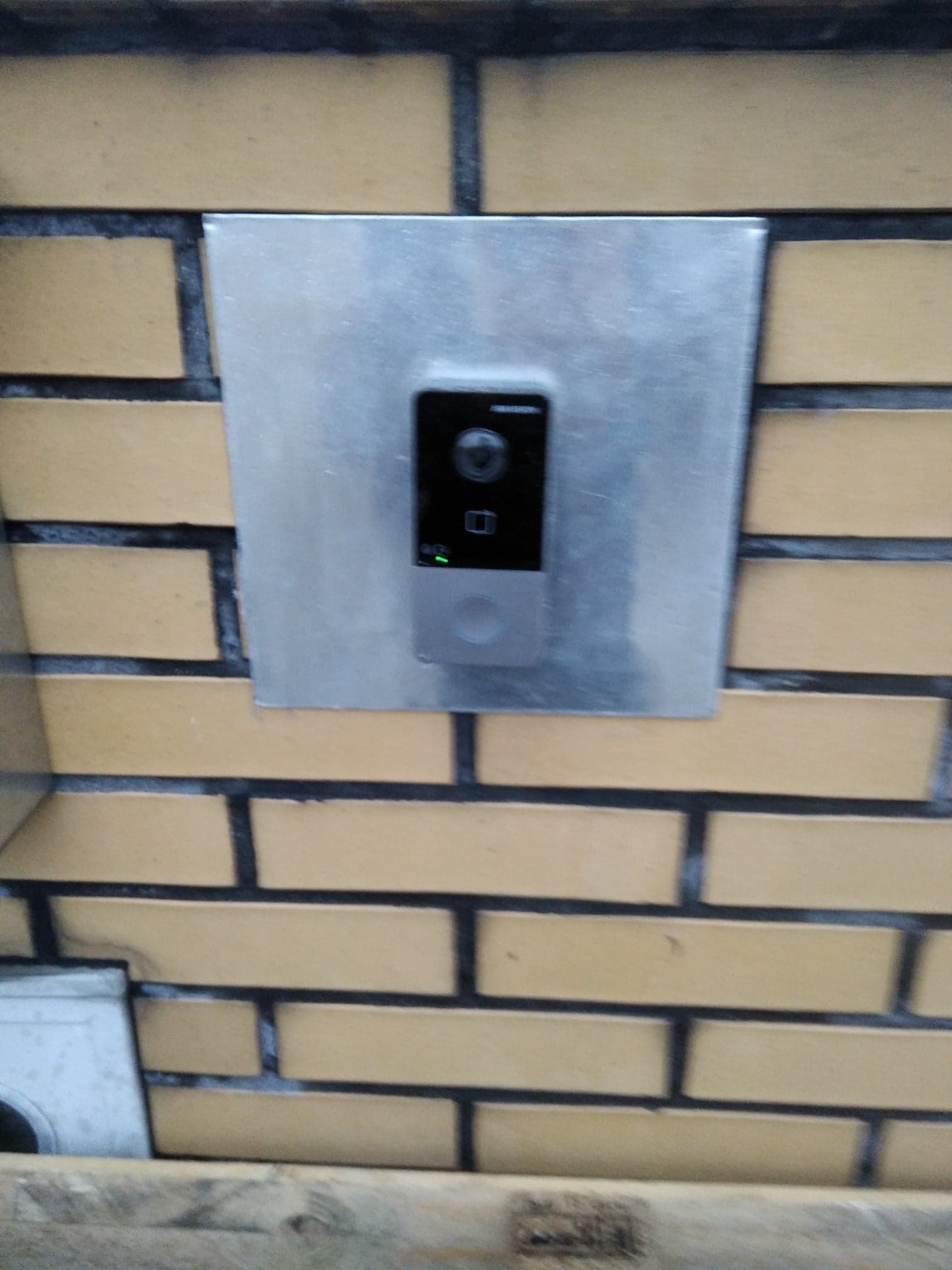 Zamontowany wideodomofon na ceglanej ścianie w Ulanowie, zabezpieczony metalową płytką. Widoczna zielona dioda sygnalizacyjna.