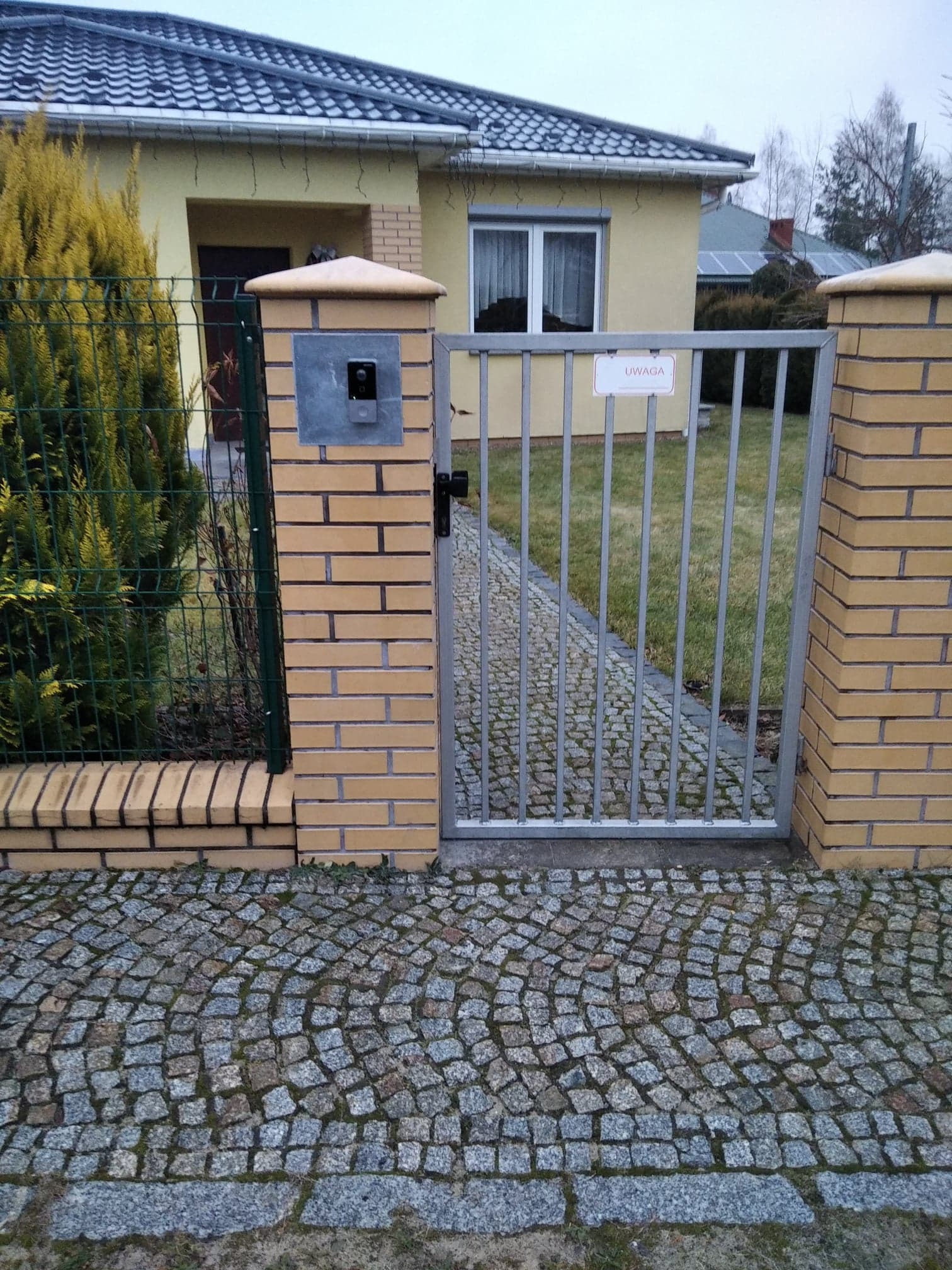 Brama wejściowa z domofonem na ceglanym słupku, tabliczka z napisem 'UWAGA' na metalowej bramie, kostka brukowa na podjeździe.