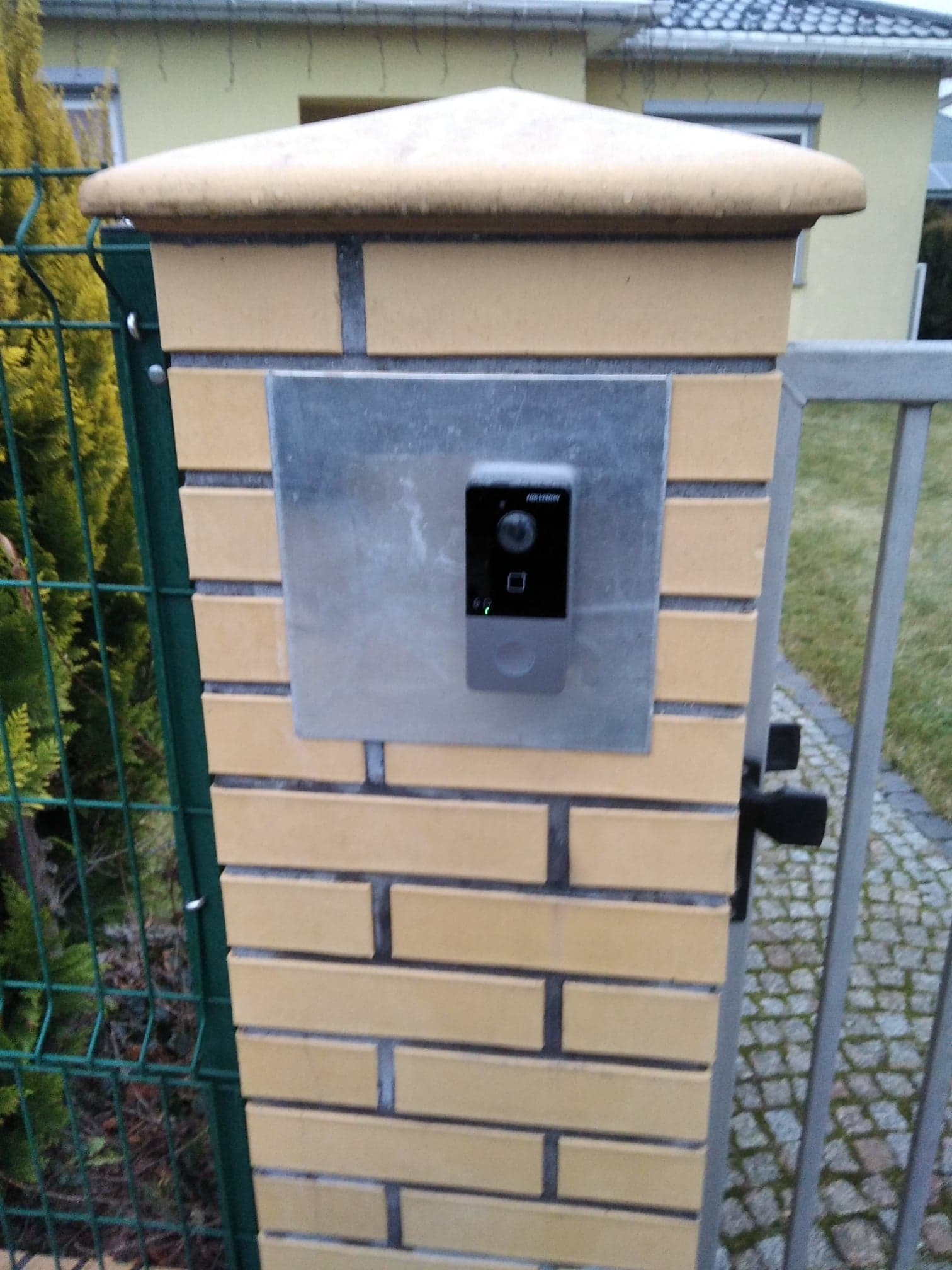 Domofon Hikvision zamontowany na metalowej płycie, przymocowanej do słupka z cegły klinkierowej przy furtce.