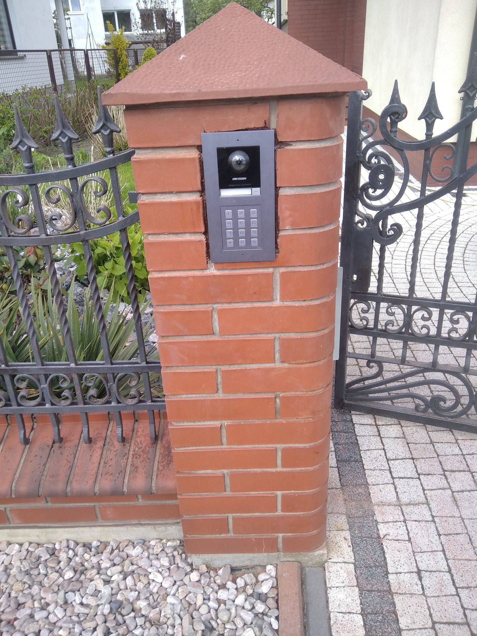 Domofon z kamerą Hikvision zamontowany na ceglanym słupku ogrodzenia z kutą bramą i ścieżką z kostki brukowej.