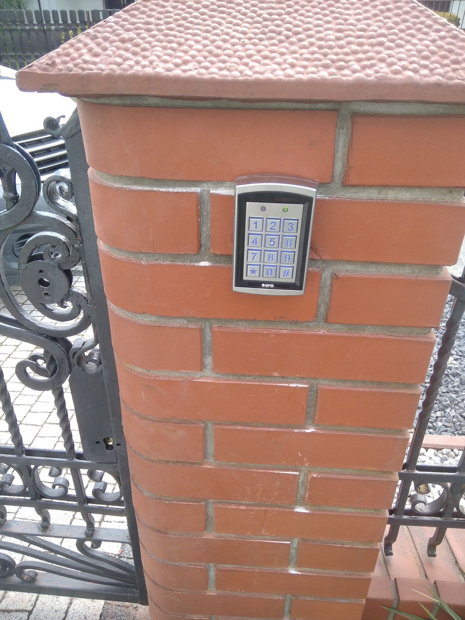 Klawiatura numeryczna systemu kontroli dostępu zamontowana na ceglanym słupku bramy wjazdowej z ozdobnym, kutym metalem.