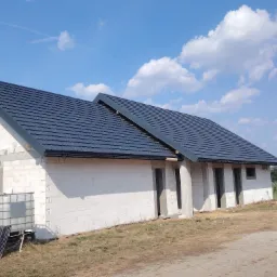 Niedokończony budynek z białych bloczków z nowym, ciemnym dachem z dachówki na tle błękitnego nieba z chmurami, widoczne rusztowanie i zbiornik na wodę.