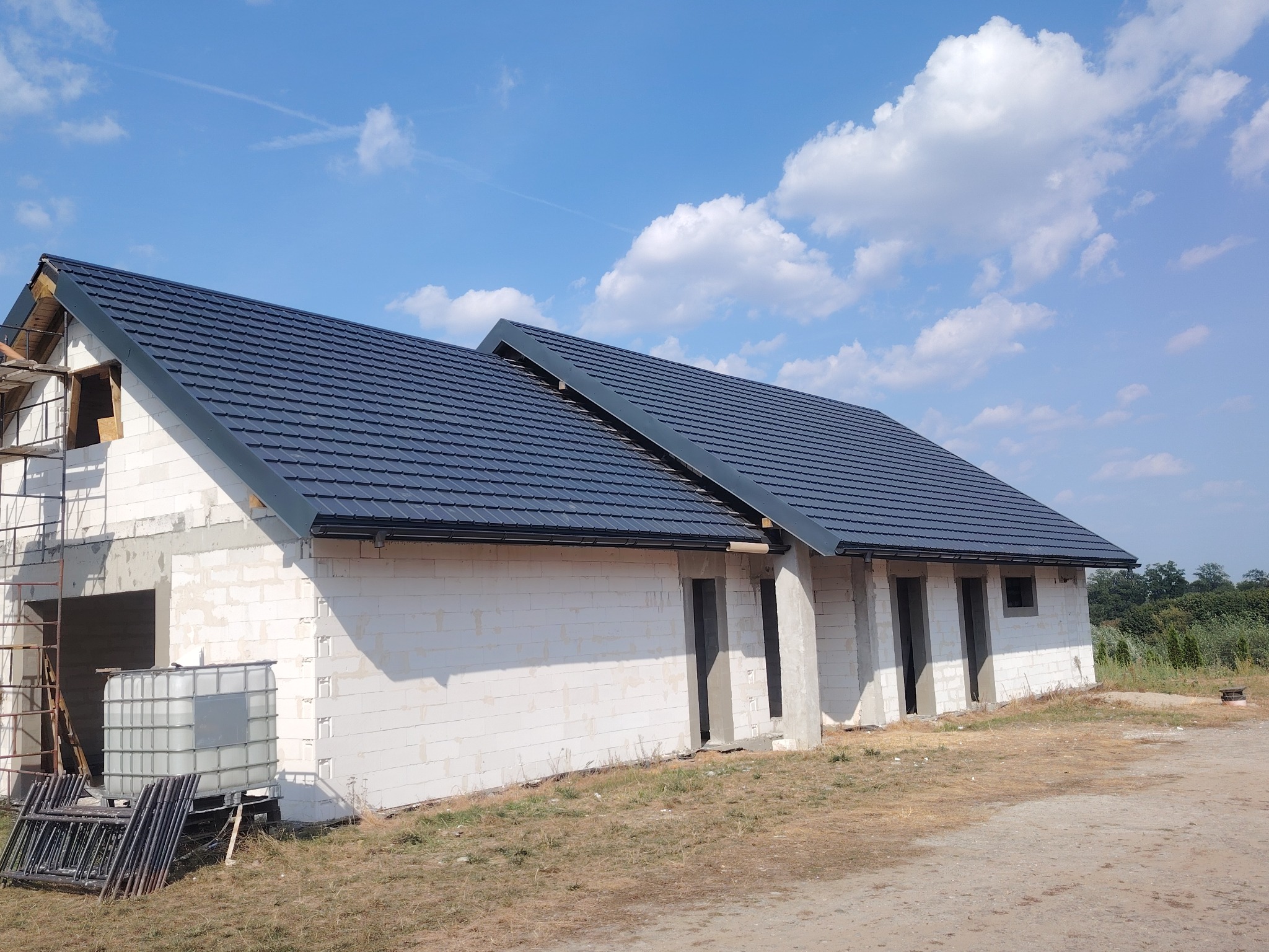 Niedokończony budynek z białych bloczków z nowym, ciemnym dachem z dachówki na tle błękitnego nieba z chmurami, widoczne rusztowanie i zbiornik na wodę.