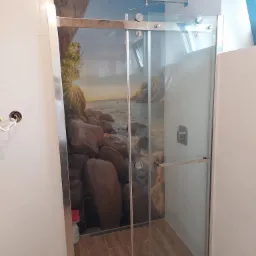 Szklana kabina prysznicowa z chromowanymi okuciami i grafiką plaży z kamieniami na ścianie, zamontowana w wykończonej łazience z białymi płytkami ściennymi i brązowymi płytkami podłogowymi...