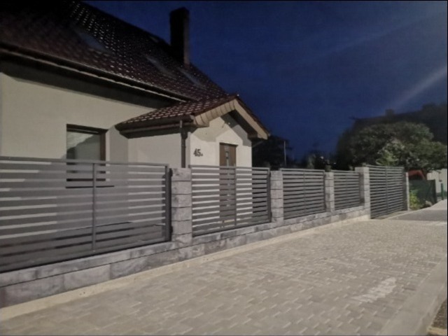 Nowoczesne ogrodzenie panelowe w kolorze szarym z betonowymi słupkami, zamontowane przed domem z numerem 45, widoczne kostka brukowa i ciemne niebo.