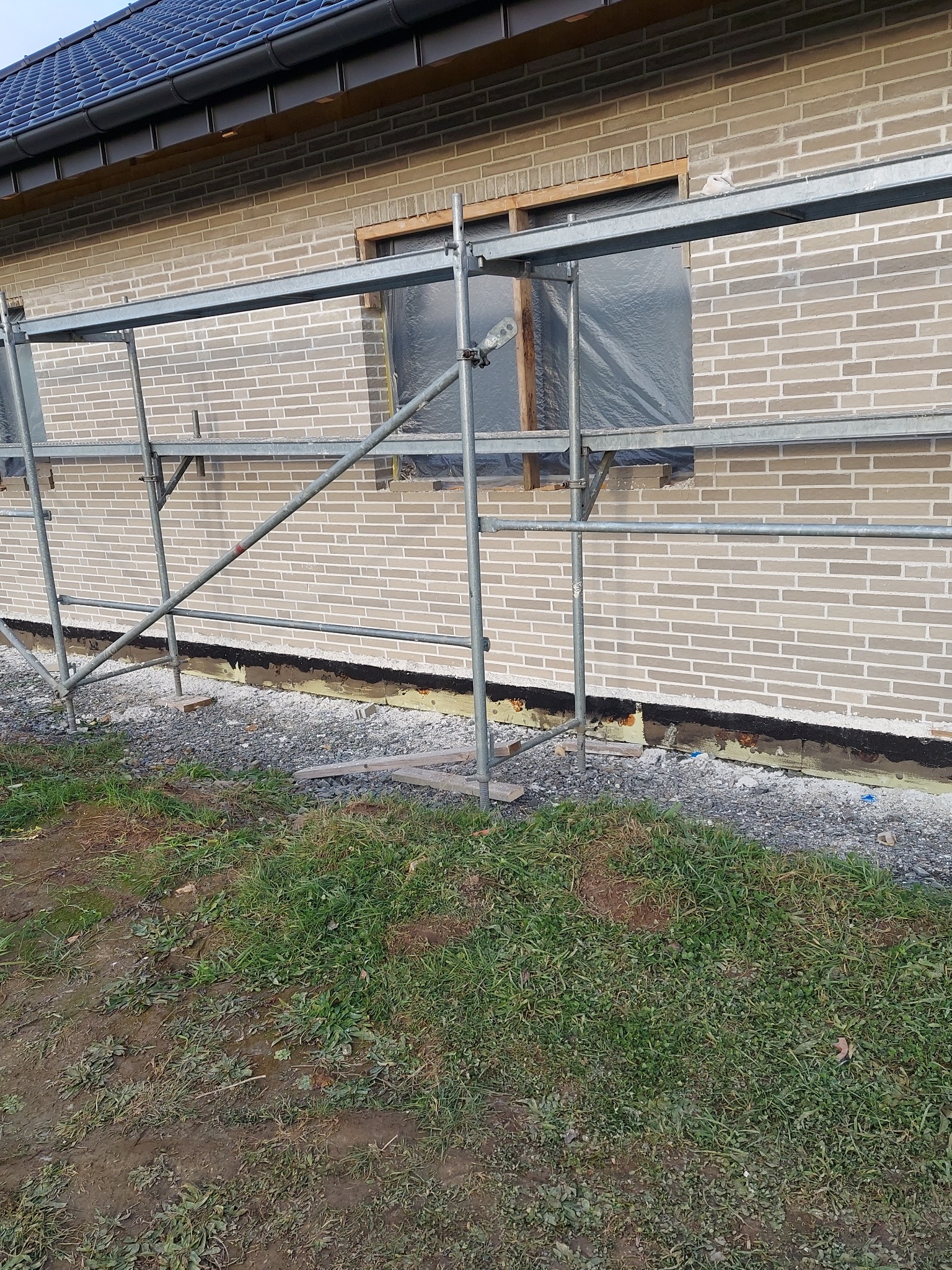 Elewacja budynku z jasnej cegły, okno zabezpieczone folią, widoczne rusztowanie i przygotowane podłoże z izolacją przy gruncie.