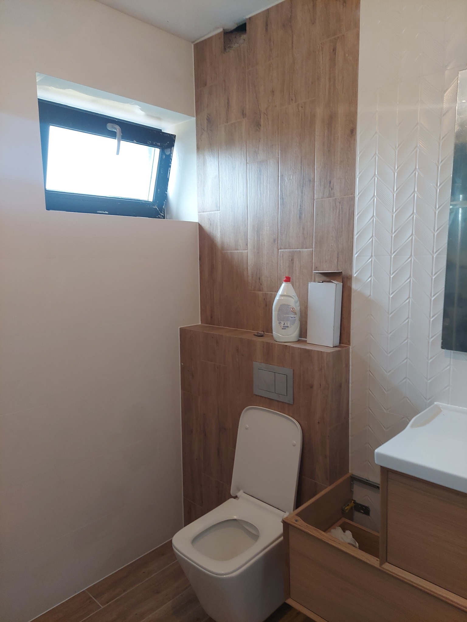 Niedokończona łazienka z białą muszlą klozetową, umywalką z drewnianą szafką bez drzwi, brązowymi płytkami imitującymi drewno oraz białymi płytkami w jodełkę, małym oknem i butelką płynu...