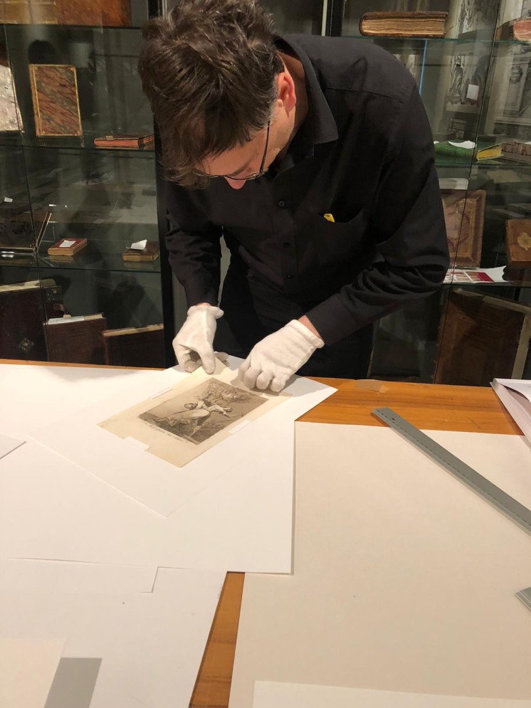 Mężczyzna w czarnej koszuli i białych rękawiczkach delikatnie obchodzi się z akwafortą z cyklu Los Caprichos Francisco Goya, leżącą na białym papierze, na drewnianym stole, w tle gabloty...