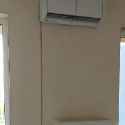 Biały klimatyzator marki AUX, jeszcze z folią ochronną, zamontowany na beżowej ścianie w pokoju, grzejnik zabezpieczony folią malarską.