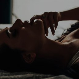 katrynphoto - Kobieta leżąca na plecach w łóżku, z ręką przy ustach, w stonowanej kolorystyce.