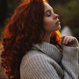 katrynphoto - Portret kobiety z rudymi, kręconymi włosami w profilu, z zamkniętymi oczami, ubrana w gruby, szary sweter, oświetlona ciepłym, naturalnym światłem.