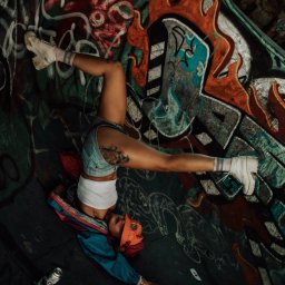 katrynphoto - Kobieta w dżinsowych szortach i białym topie wykonuje stójkę na tle ściany pokrytej kolorowymi graffiti, z widocznymi puszkami po farbie i biletami leżącymi na ziemi.