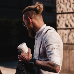 katrynphoto - Mężczyzna z brodą i fryzurą w kok, w okularach, ubrany w białą koszulę na szelkach, trzymający kubek Starbucks, z przypinkami na skórzanej kurtce.