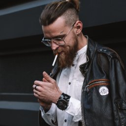 katrynphoto - Mężczyzna z brodą i tatuażem 'ART' nad brwią, w okularach, białej koszuli, skórzanej kurtce i z zegarkiem na ręku, odpala papierosa zapalniczką.