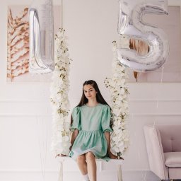 katrynphoto - Nastolatka w miętowej sukience siedzi na huśtawce ozdobionej białymi kwiatami, z balonami w kształcie cyfr '1' i '5' po bokach, w jasnym wnętrzu z marmurową podłogą i różowym fotelem w tle.