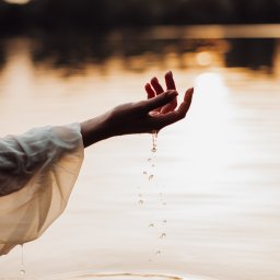 katrynphoto - Dłoń kobiety w zwiewnej białej szacie unosi się nad wodą, z palców spływają krople, tworząc delikatne fale na powierzchni jeziora w ciepłym, zachodzącym słońcu.