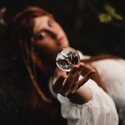 katrynphoto - Portret kobiety z rudymi włosami, trzymającej w dłoni szklaną kulę, w której odbija się otoczenie, ubrana w białą bluzkę z bufiastymi rękawami, z delikatnym wiankiem we włosach, na rozmytym...