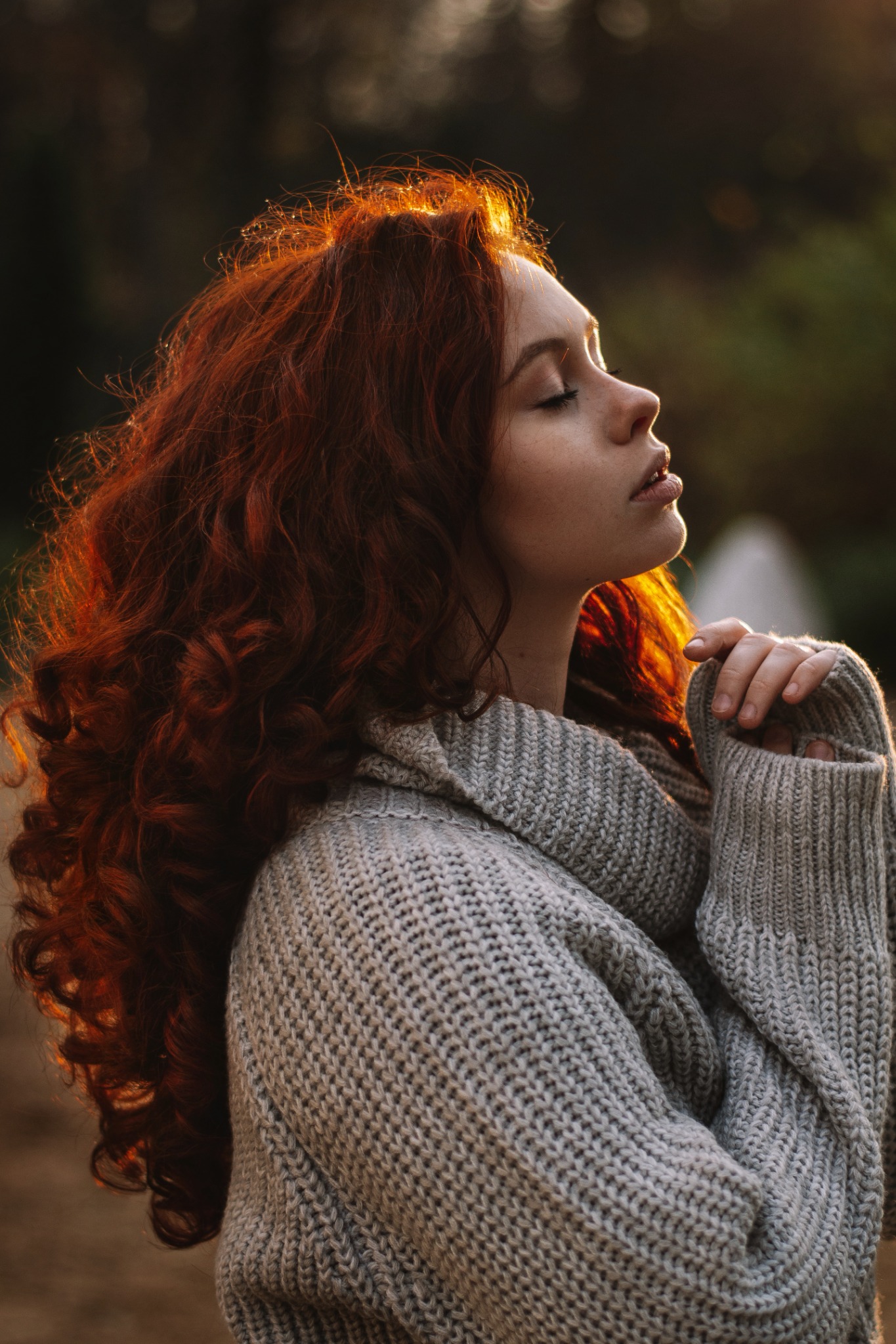 Portret kobiety z rudymi, kręconymi włosami w profilu, z zamkniętymi oczami, ubrana w gruby, szary sweter, oświetlona ciepłym, naturalnym światłem.