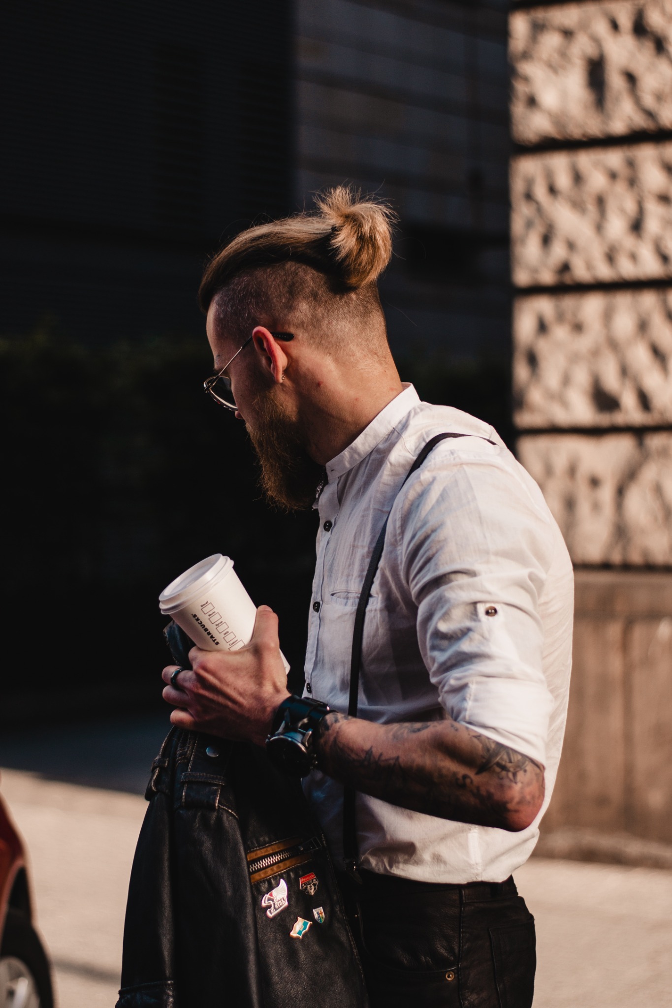 Mężczyzna z brodą i fryzurą w kok, w okularach, ubrany w białą koszulę na szelkach, trzymający kubek Starbucks, z przypinkami na skórzanej kurtce.