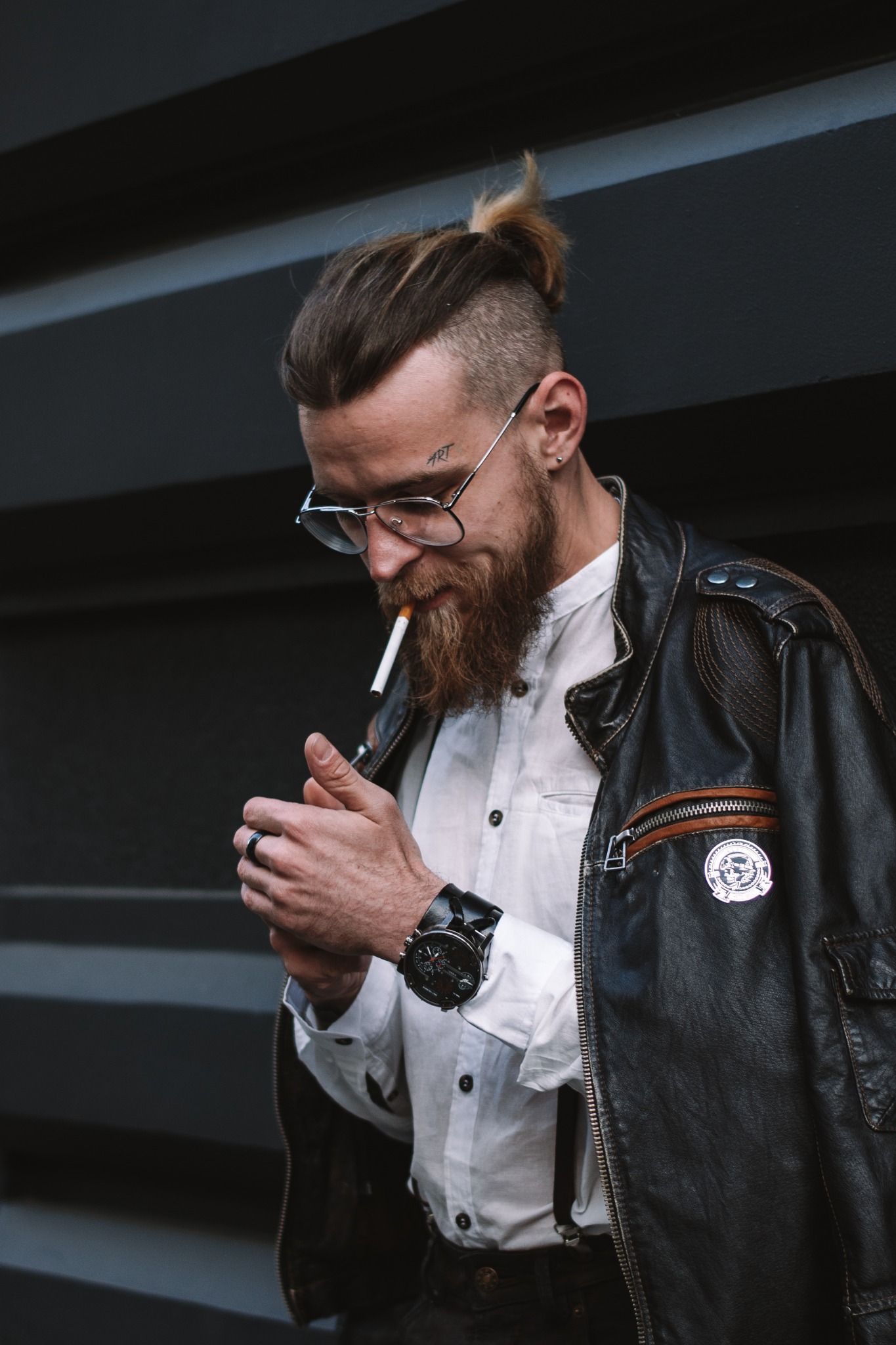 Mężczyzna z brodą i tatuażem 'ART' nad brwią, w okularach, białej koszuli, skórzanej kurtce i z zegarkiem na ręku, odpala papierosa zapalniczką.