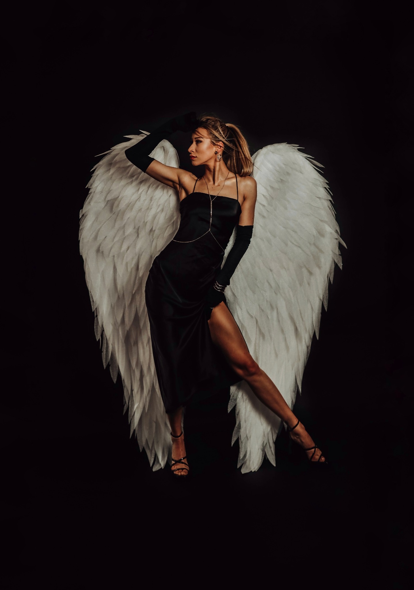 Kobieta w czarnej sukience i długich rękawiczkach pozuje z białymi skrzydłami na czarnym tle, długa złota biżuteria na ciele, włosy spięte w kucyk.
