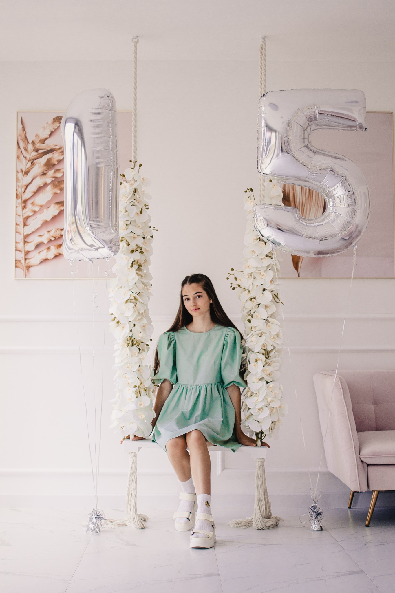 Nastolatka w miętowej sukience siedzi na huśtawce ozdobionej białymi kwiatami, z balonami w kształcie cyfr '1' i '5' po bokach, w jasnym wnętrzu z marmurową podłogą i różowym fotelem w tle.