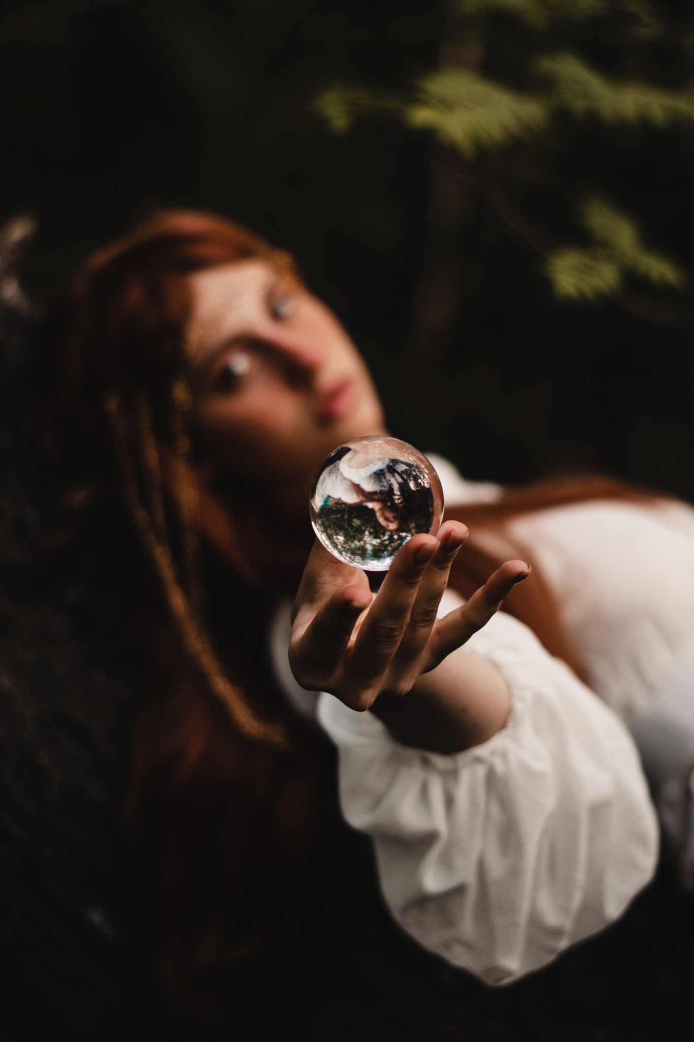 Portret kobiety z rudymi włosami, trzymającej w dłoni szklaną kulę, w której odbija się otoczenie, ubrana w białą bluzkę z bufiastymi rękawami, z delikatnym wiankiem we włosach, na rozmytym...