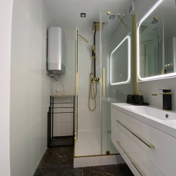 DomPRO Patryk Dąbrowski - Nowoczesna łazienka z kabiną prysznicową typu walk-in, czarno-złotą armaturą, podświetlanym lustrem i ciemną podłogą imitującą marmur oraz srebrnym bojlerem.