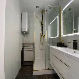 Nowoczesna łazienka z kabiną prysznicową typu walk-in, czarno-złotą armaturą, podświetlanym lustrem i ciemną podłogą imitującą marmur oraz srebrnym bojlerem.