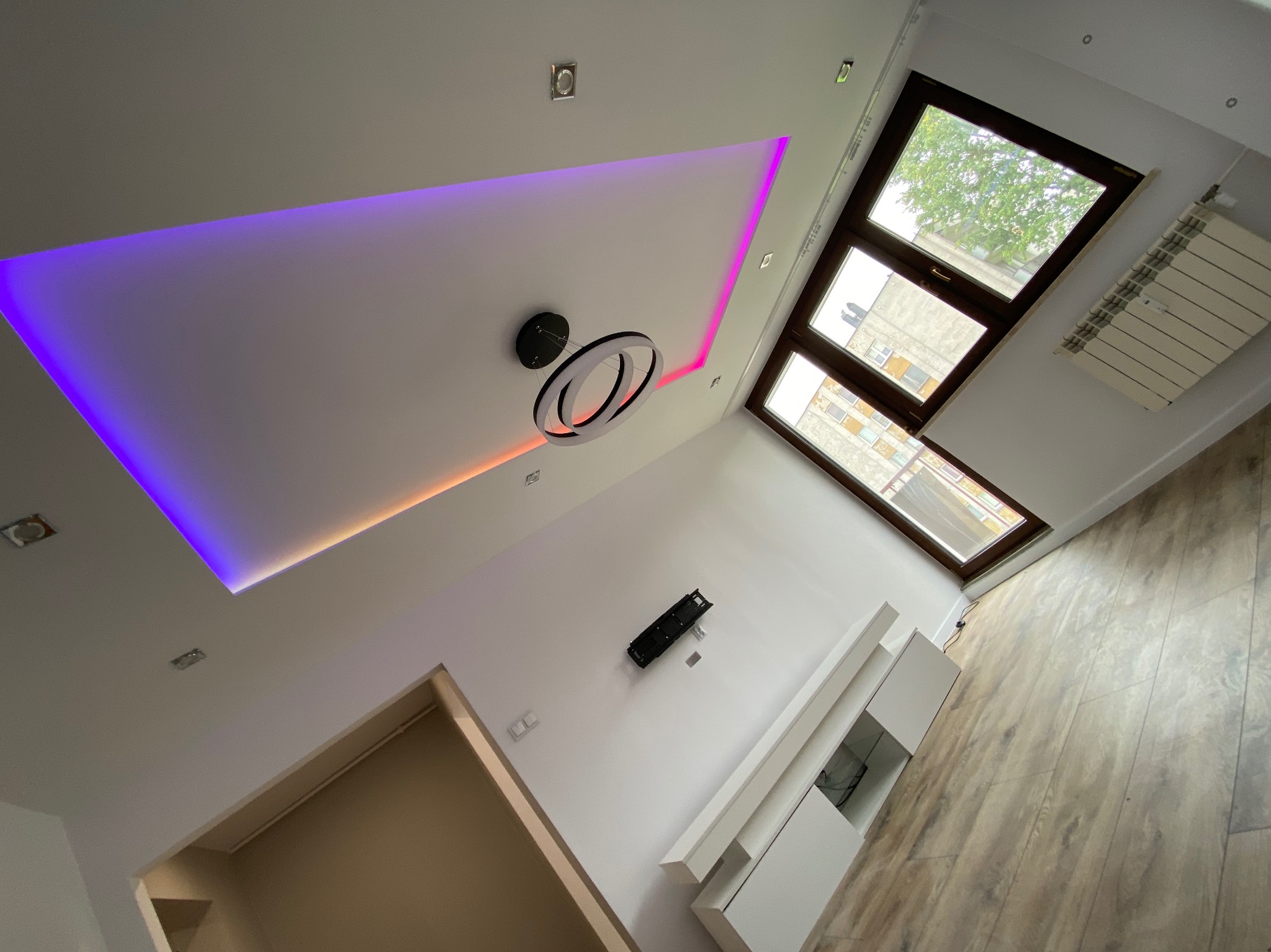 Nowoczesny salon z podświetlanym sufitem podwieszanym w kolorach fioletu i pomarańczy, okrągła lampa sufitowa z trzema pierścieniami, drewniana podłoga i białe meble.