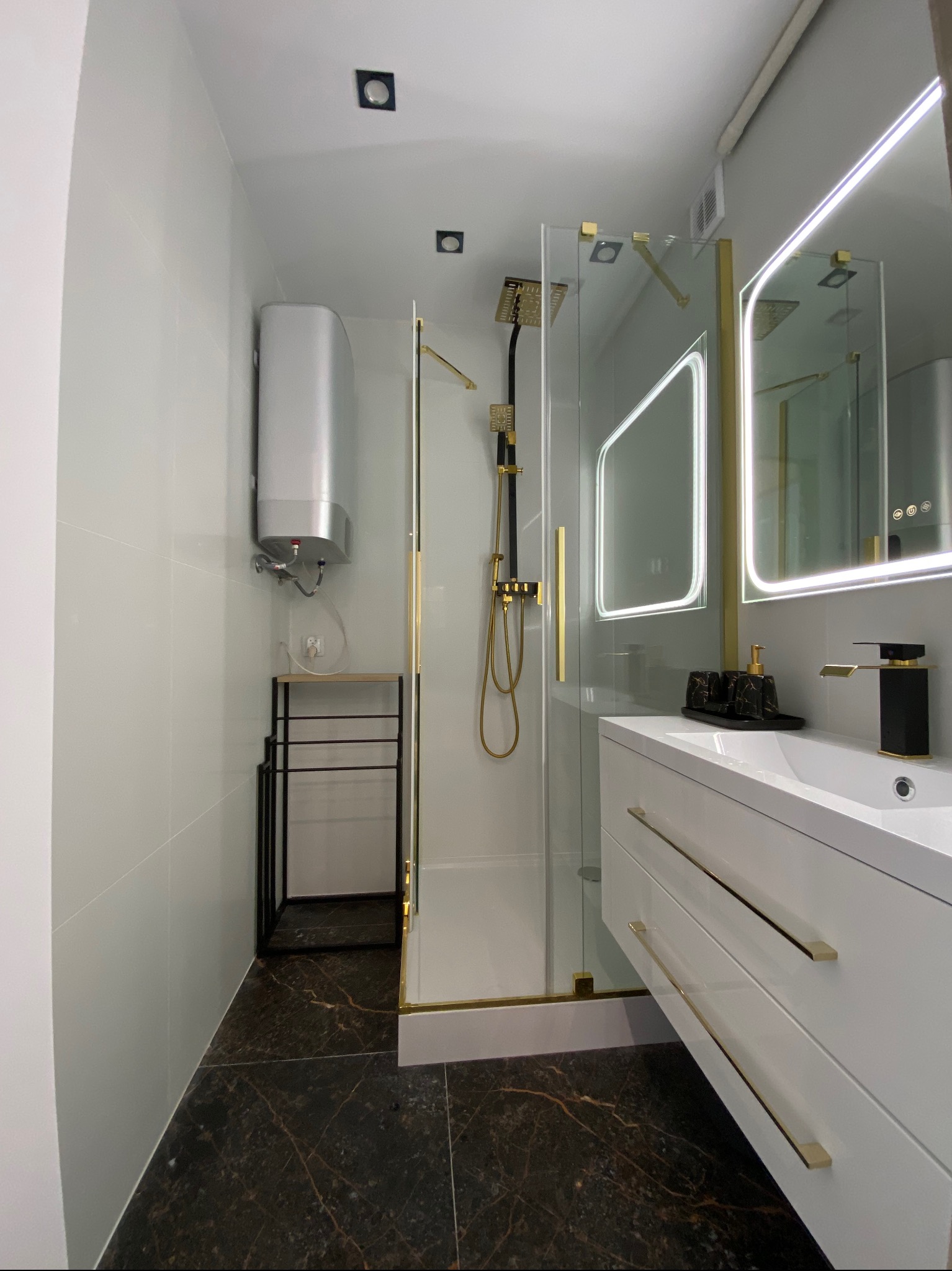 Nowoczesna łazienka z kabiną prysznicową typu walk-in, czarno-złotą armaturą, podświetlanym lustrem i ciemną podłogą imitującą marmur oraz srebrnym bojlerem.