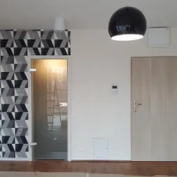 Wnętrze mieszkania po remoncie: ściana z geometryczną tapetą w odcieniach szarości, szklane drzwi z widoczną drabiną w tle, drewniane drzwi wejściowe, czarna lampa wisząca i białe ściany.