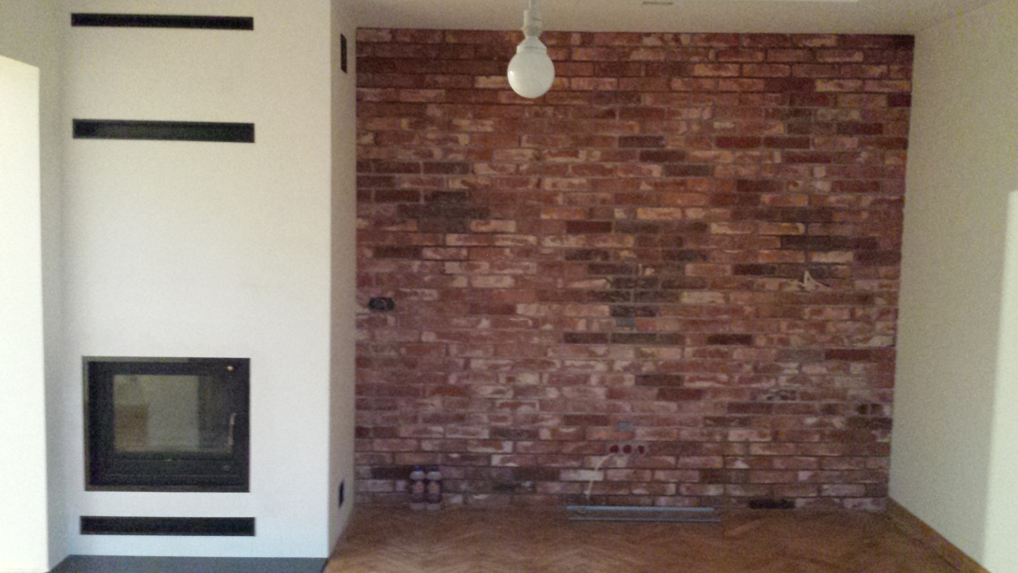 Surowa ściana z czerwonej cegły z widocznymi przewodami elektrycznymi i żarówką w minimalistycznym wnętrzu z podłogą w jodełkę i nowoczesnym kominkiem.