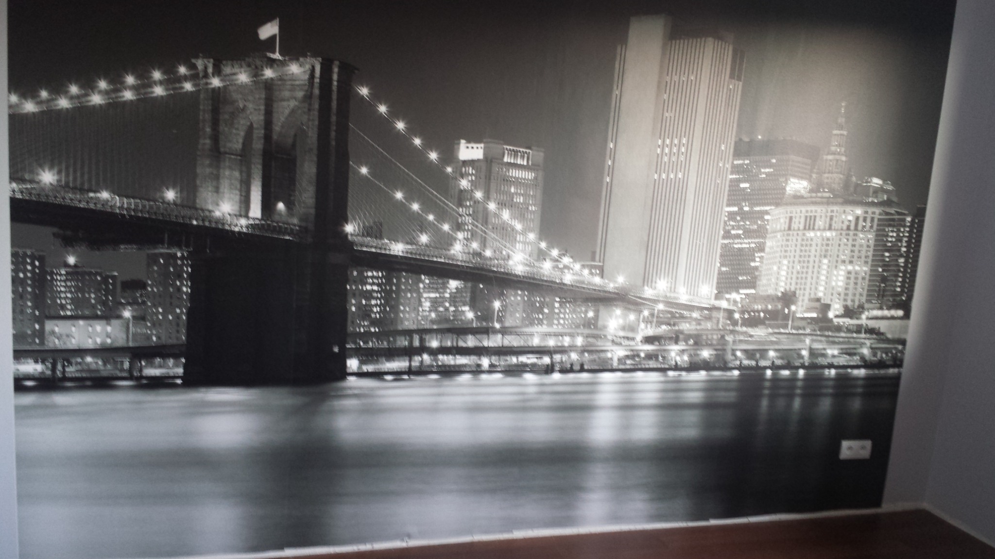 Fototapeta przedstawiająca nocny widok na most Brooklyński i panoramę Manhattanu w czerni i bieli, umieszczona na ścianie w pomieszczeniu z widocznym gniazdkiem elektrycznym.
