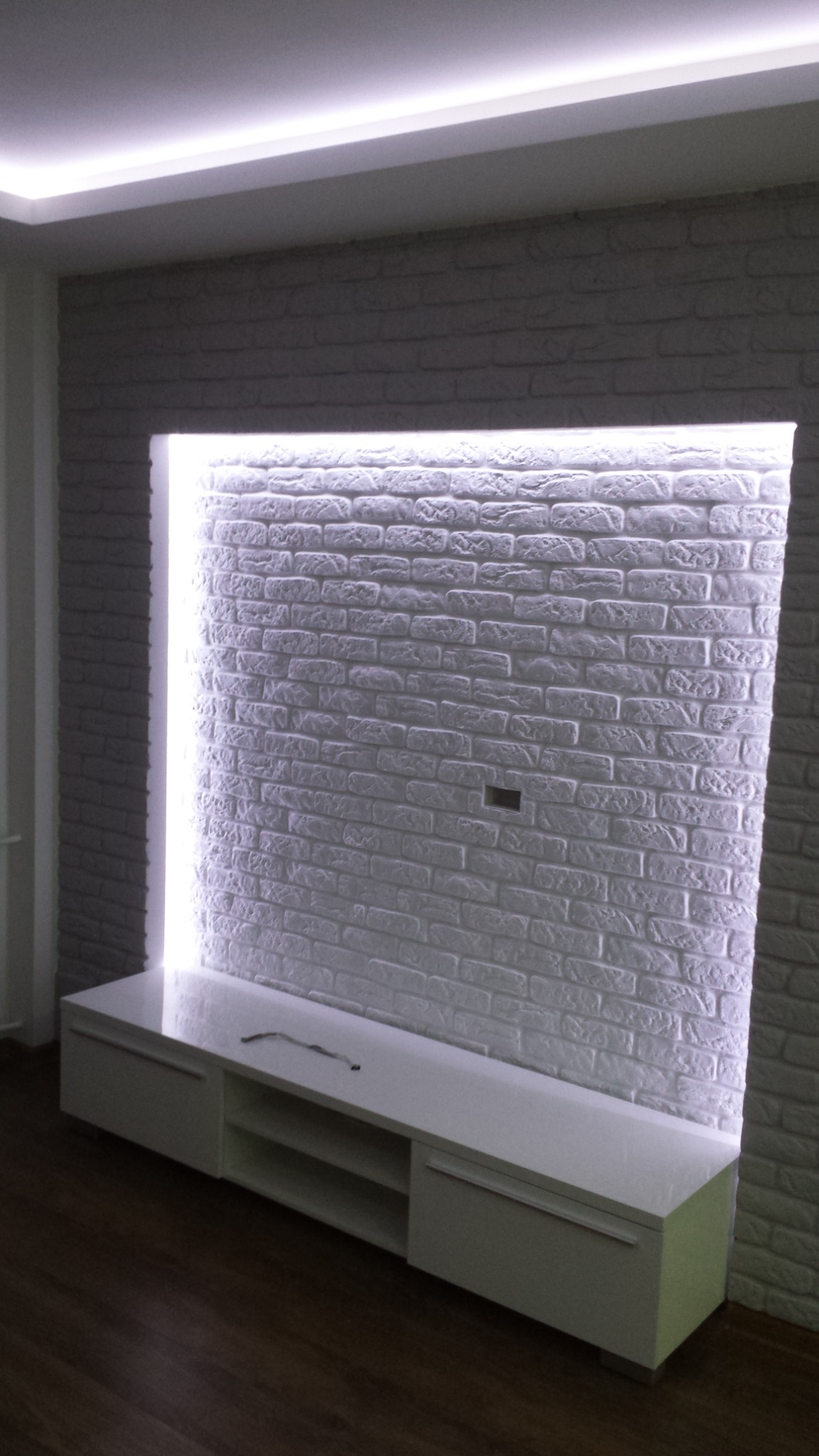 Ściana z białej cegły dekoracyjnej z podświetleniem LED i szafką RTV w nowoczesnym salonie.