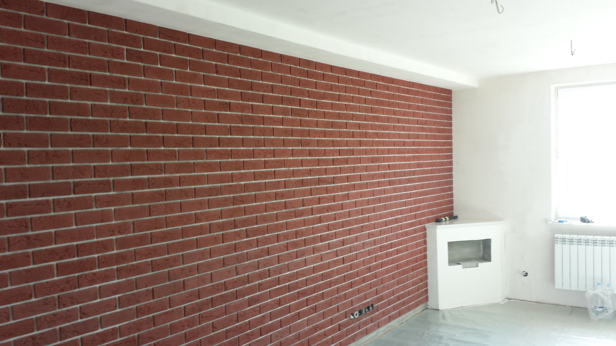 Ściana z cegły klinkierowej w nowym wnętrzu, z widocznym kominkiem i grzejnikiem pod oknem, podłoga zabezpieczona folią.