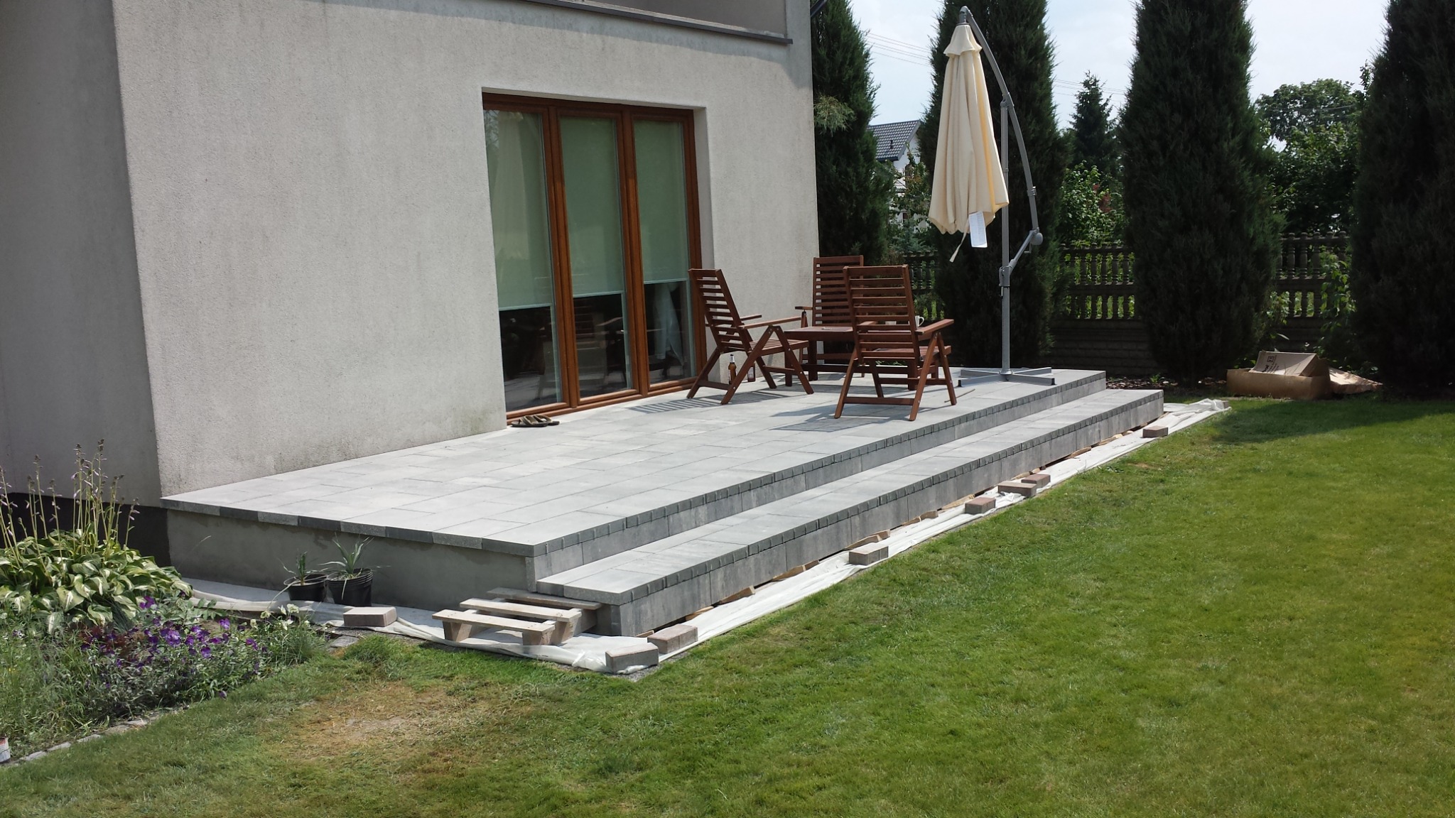 Nowoczesny taras z szarej kostki brukowej, schody wejściowe, drewniane meble ogrodowe i parasol na tle zielonego trawnika i wysokich tuj.