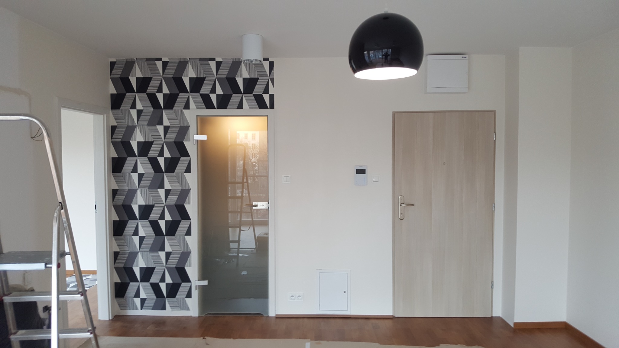 Wnętrze mieszkania po remoncie: ściana z geometryczną tapetą w odcieniach szarości, szklane drzwi z widoczną drabiną w tle, drewniane drzwi wejściowe, czarna lampa wisząca i białe ściany.