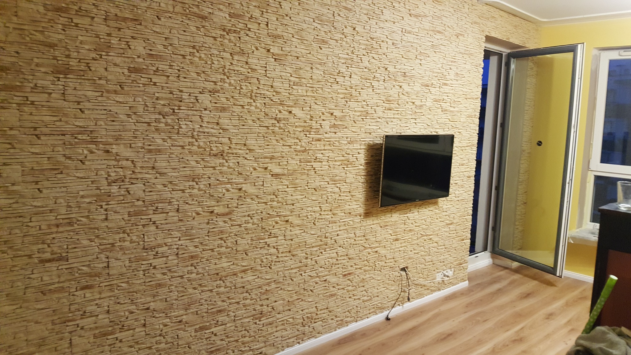 Ściana wykończona dekoracyjnym kamieniem w odcieniach beżu, z zamontowanym telewizorem, widoczne gniazdko elektryczne i fragment podłogi z paneli.