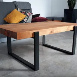 Stolik kawowy z grubym blatem z jasnego drewna i czarnymi, metalowymi nogami w kształcie litery U, na tle szarej sofy i telewizora w nowoczesnym salonie.