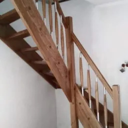 Wykończone, drewniane schody z metalowymi tralkami, prowadzące na górę, z widocznymi słojami drewna i jasnym wykończeniem, na tle białej ściany i podłogi imitującej drewno.