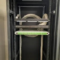 Szafa serwerowa z panelami krosowniczymi i uporządkowanymi kablami CAT6, widok z przodu na czarne wnętrze szafy z uchwytami montażowymi po bokach.