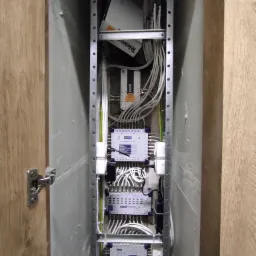 Rozdzielnia antenowa w wąskiej wnęce, widoczne multiswitche, wzmacniacz antenowy Televes Avant 6, kable koncentryczne oraz metalowa konstrukcja montażowa.